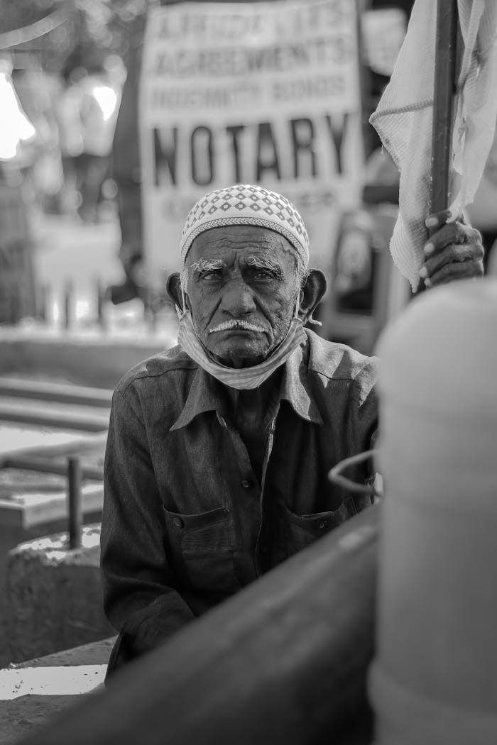 Black and white photo of an elderly man sitting near a notary sign, outdoors.