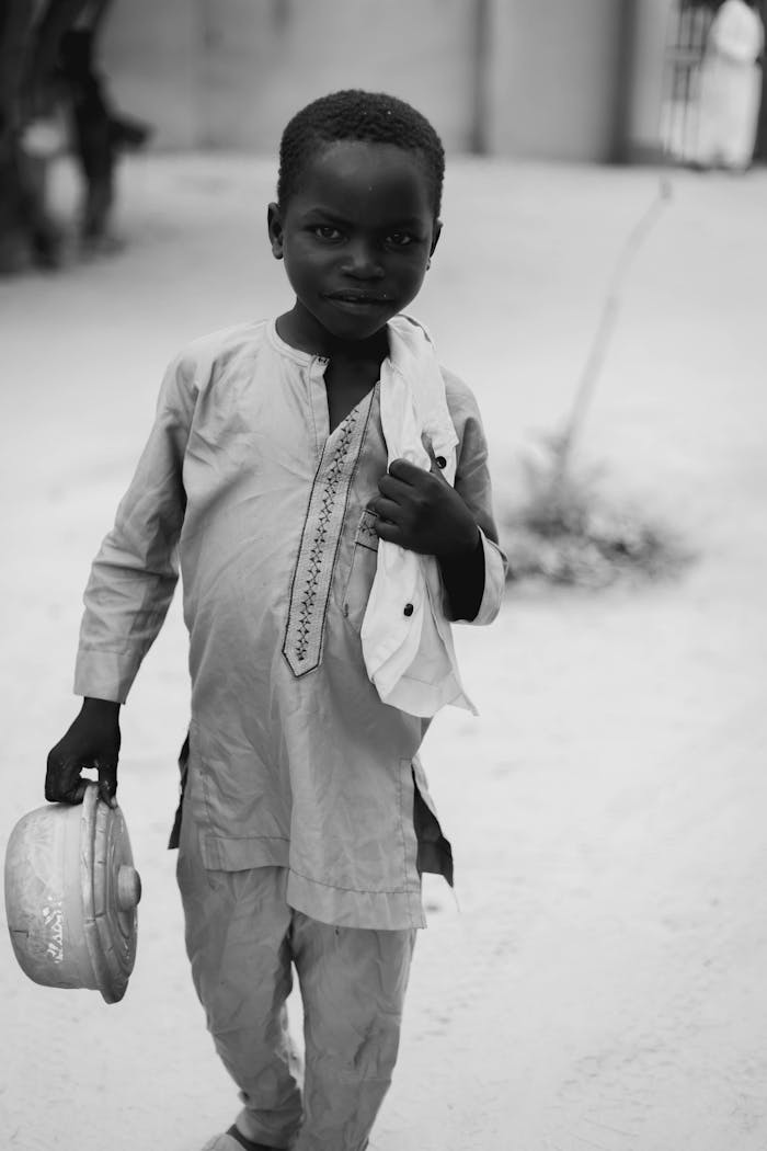 Monochrome image of a child holding a pot, dressed in traditional attire outdoors.